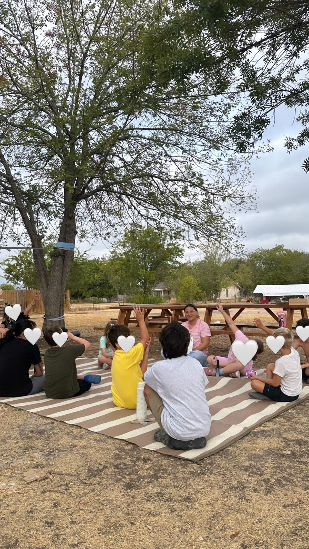 Cross-grade circle time with hands raised under campus shade